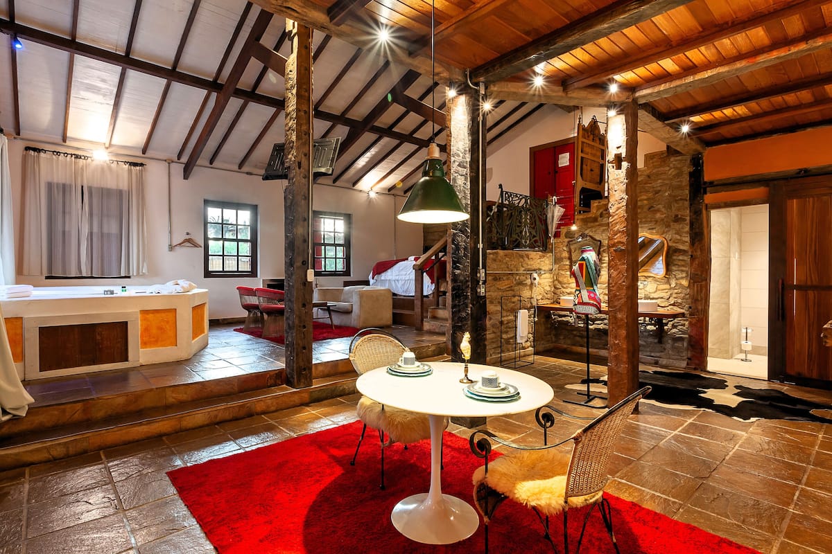 A rustic interior with a stone wall, wooden beams, a round table with chairs, and a red carpet.