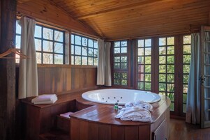 Un baño de cabaña de madera con una bañera grande, bancos de madera y ventanas amplias.