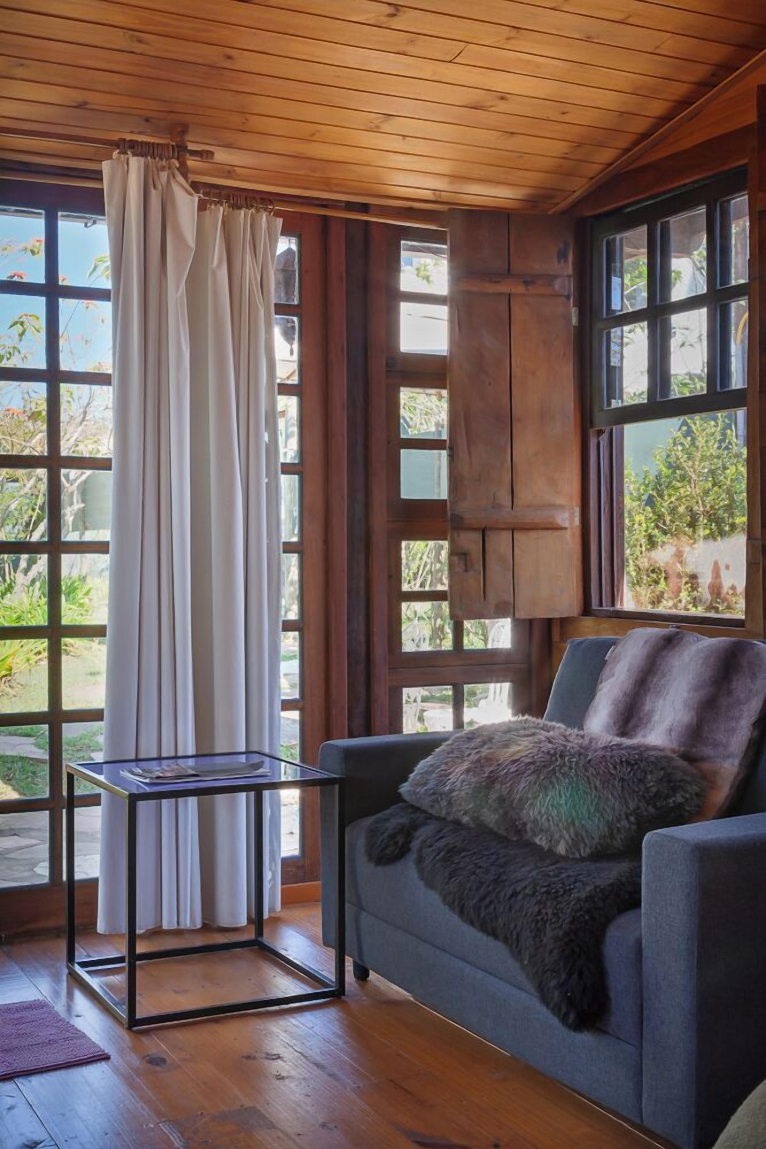 A room with wooden ceiling, a sofa with a fur throw, a glass table, and a window with curtains.