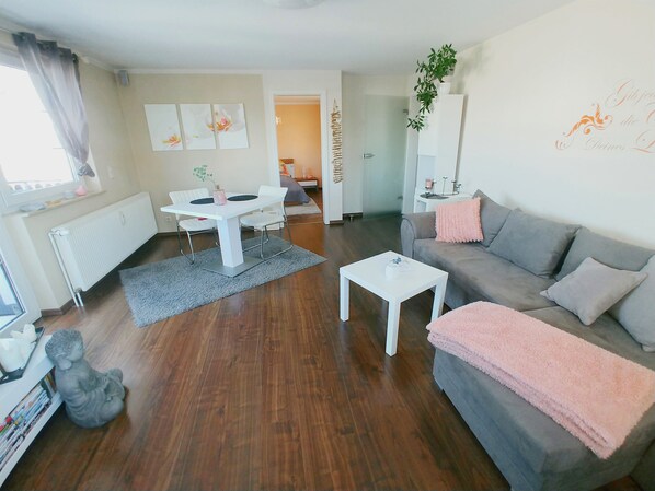 Ein modernes Wohnzimmer mit einem grauen Sofa, einem weißen Couchtisch und einem Esstisch mit Stühlen. Ein Heizkörper, ein grauer Teppich und eine kleine Statue befinden sich auf dem Boden.