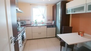 A compact kitchen with a stove, microwave, and sink. There is a window with curtains, a table with a chair, and cabinets above the counter. - Feel-good time in the Zittau Mountains (Eichgraben)