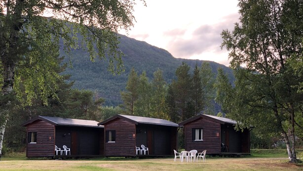 Tre cabine di legno con posti a sedere all'aperto, immerse in una zona erbosa con alberi e una montagna sullo sfondo.
