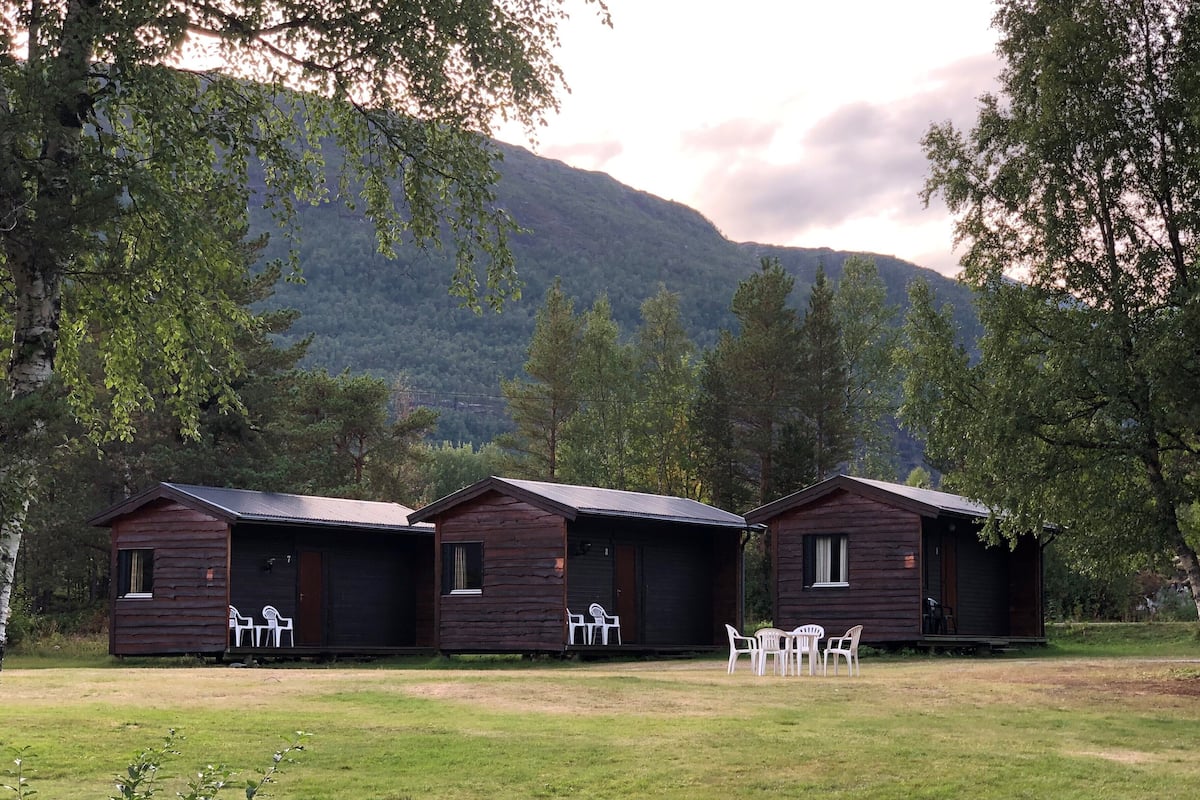 Three wooden cabins with outdoor seating, set in a grassy area with trees and a mountain in the background.