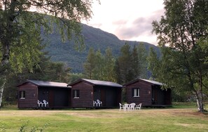 Three wooden cabins with outdoor seating, set in a grassy area with trees and a mountain in the background. - Tømmerneset Camping (Hamaroy)