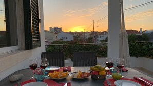 Une table dressée pour un repas, avec des assiettes, des verres et un saladier de fruits, sur un balcon donnant sur un coucher de soleil.