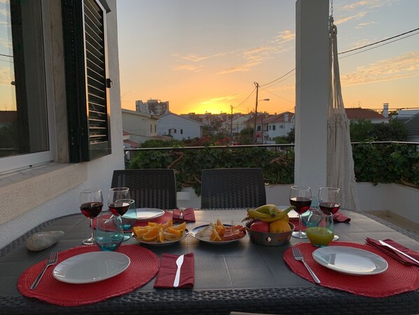 A table set for a meal with plates, glasses, and a fruit bowl on a balcony overlooking a sunset.