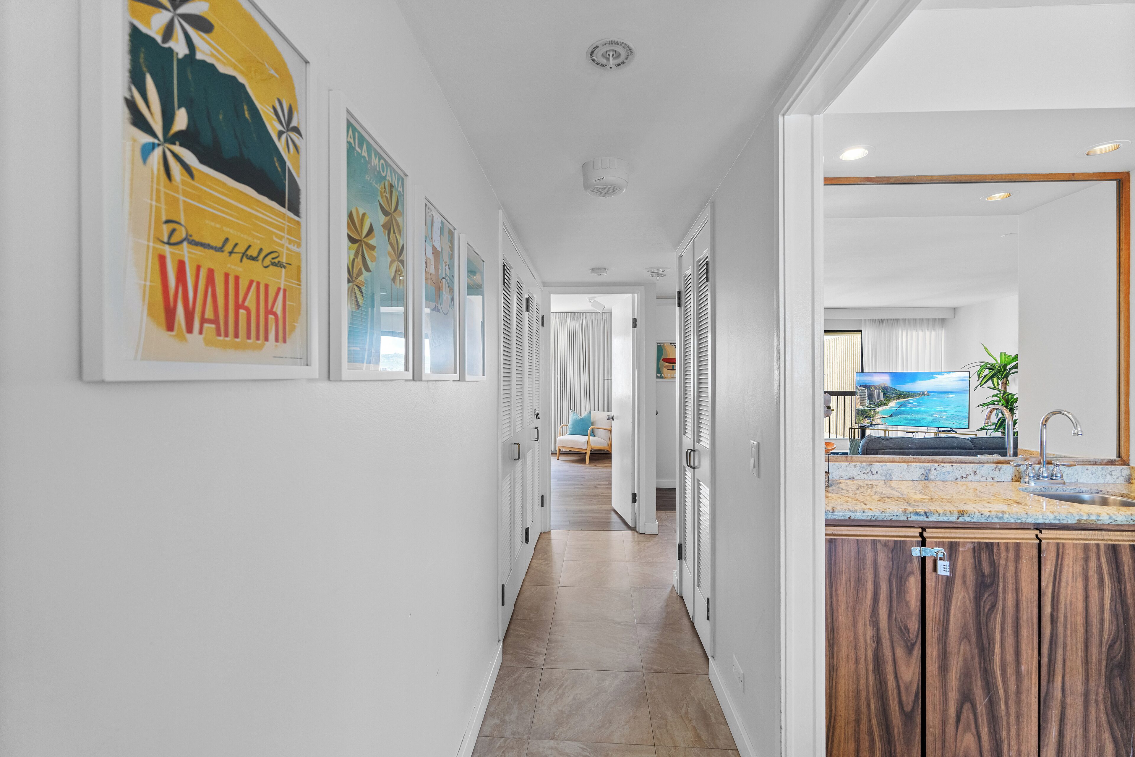 A hallway with framed artwork, a kitchen area with a sink and countertop, and a living room with a couch and TV.