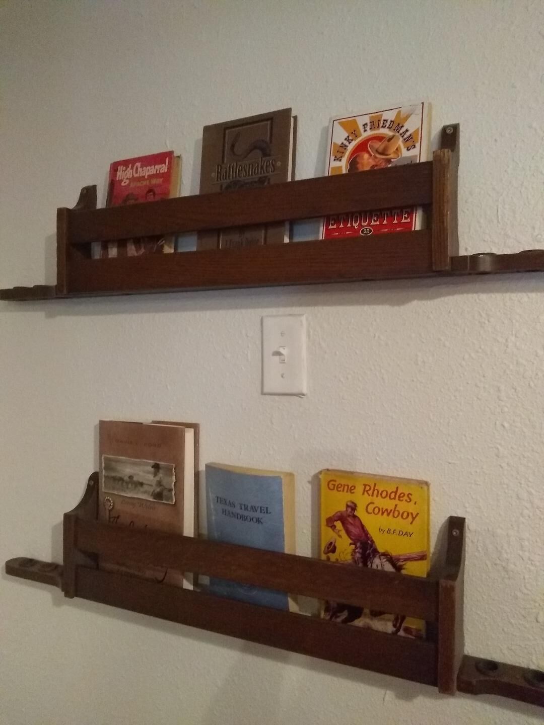 Two wooden shelves mounted on a wall, each holding various books and a cigarette pack.
