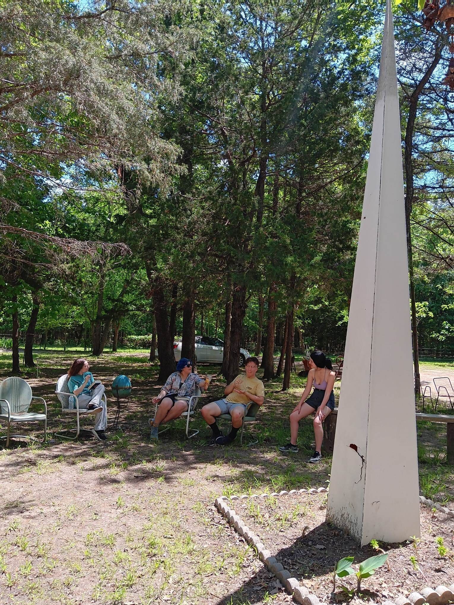 Ein Park mit einem weißen Denkmal, Bäumen und Personen, die auf Stühlen sitzen.
