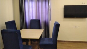 A room with a table and four chairs, an air conditioning unit, a wall-mounted TV, and a window with curtains.