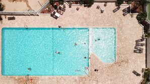 A large rectangular swimming pool with clear blue water, surrounded by a sandy beach area with lounge chairs and umbrellas.
