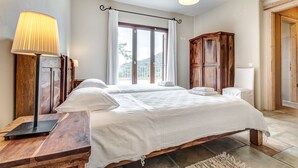 A bedroom with a large bed, wooden headboard, two bedside lamps, a wooden wardrobe, and a window with curtains. - Mastrogiannis villa Myrtia (Kavvadades)