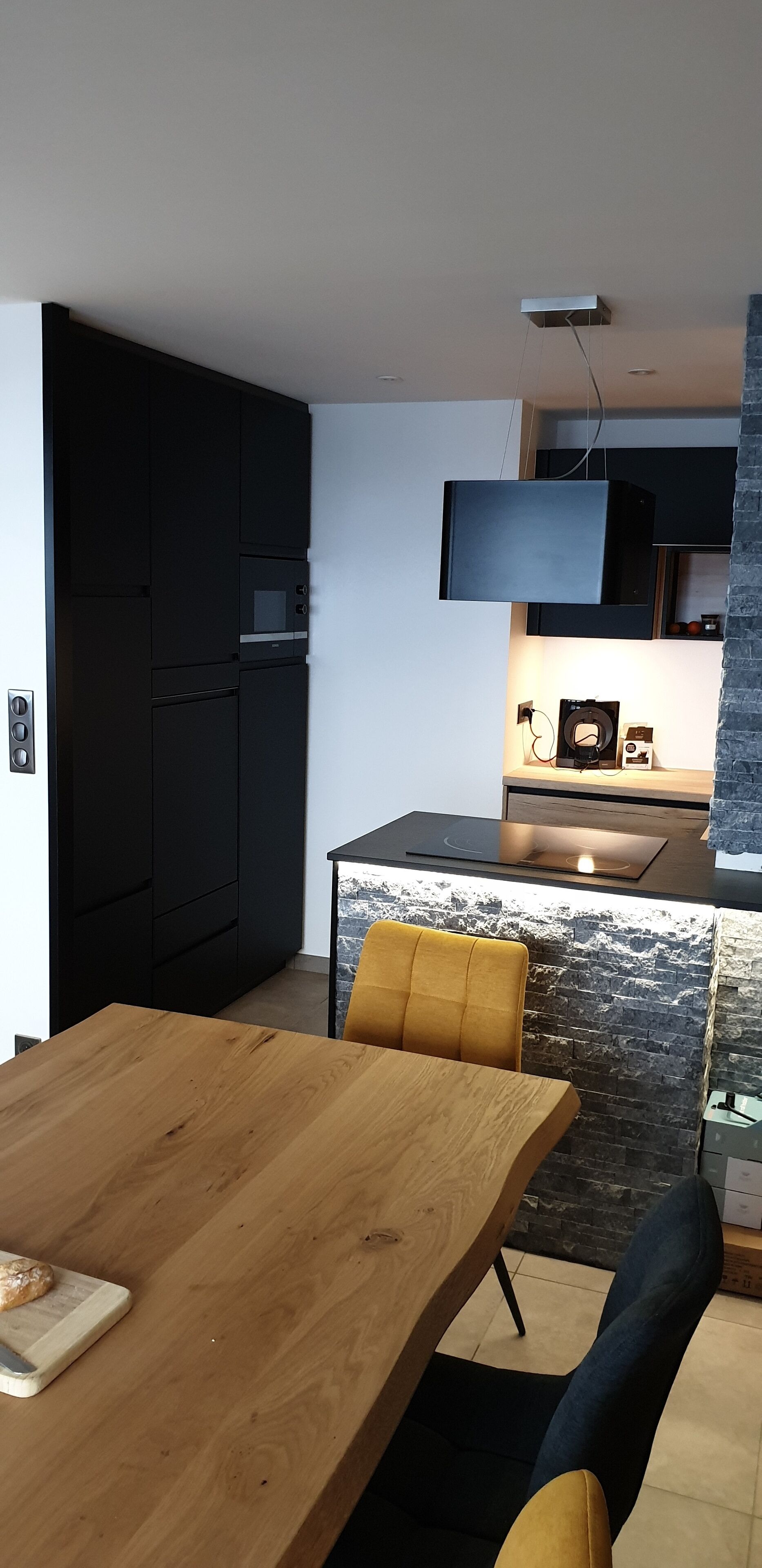 A modern kitchen with built-in appliances, a dining area with a wooden table and yellow chairs, and a stone accent wall.