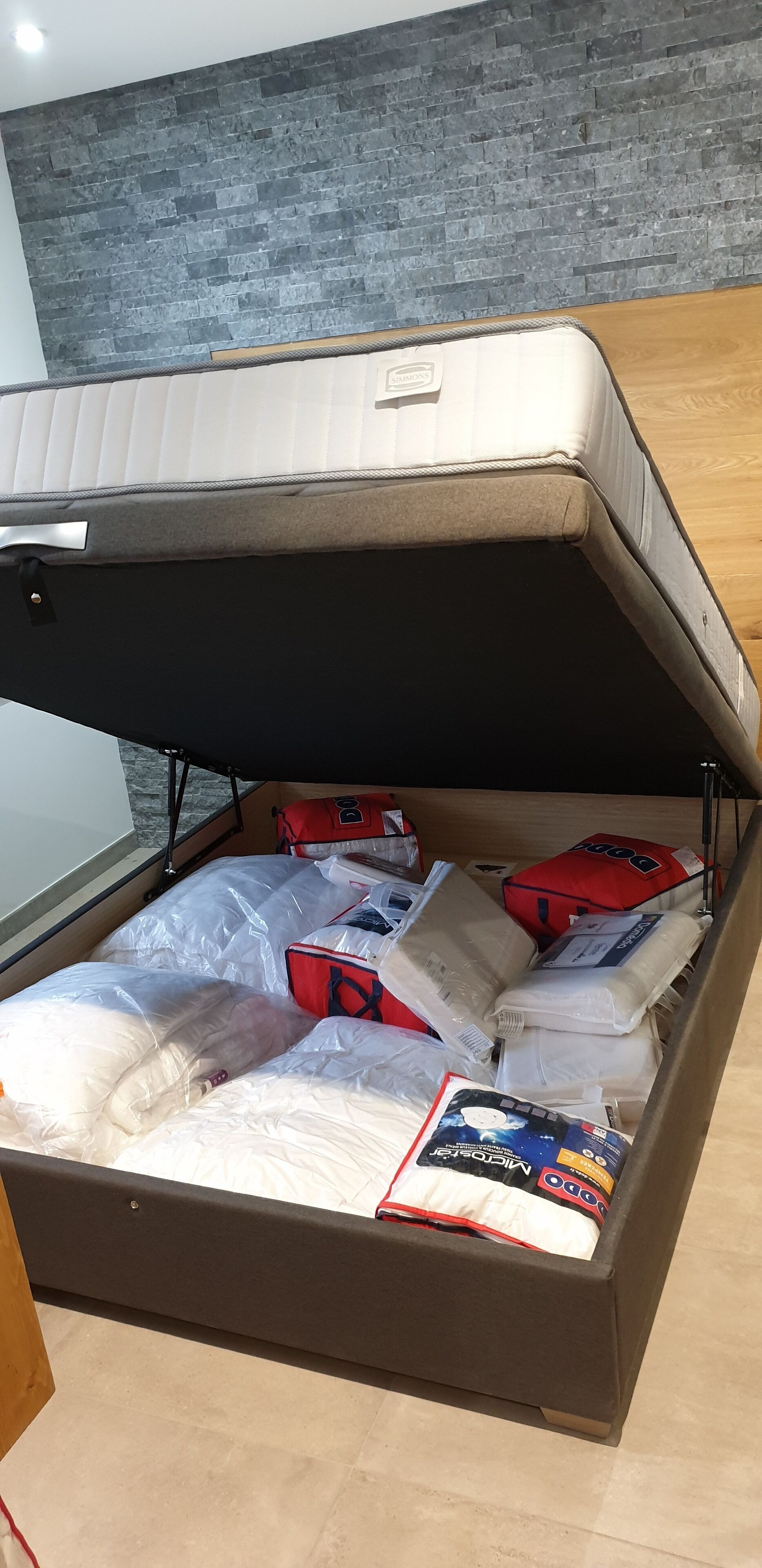 A bed with a storage compartment underneath, containing various items like pillows, bags, and a box.
