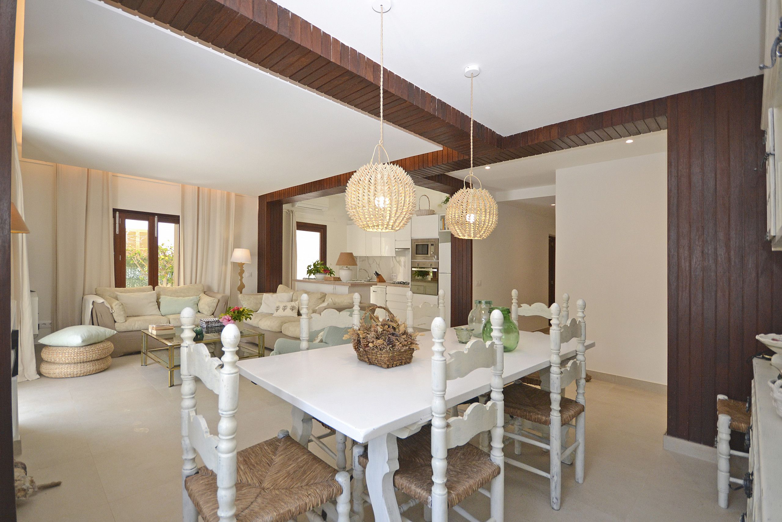A dining area with a white table and chairs, a hanging light fixture, and a view into the kitchen.