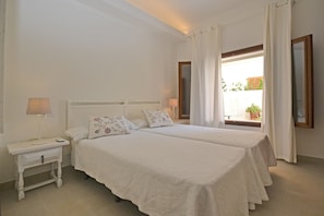A double bed with floral pillows, a bedside table with a lamp, and a window with curtains.