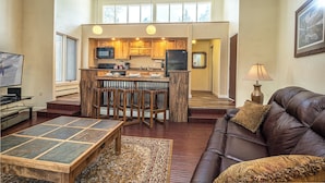 A spacious living area with a large brown leather sofa, a wooden coffee table, and a patterned rug. A kitchen with wooden cabinets and a bar area is visible in the background. - Gold Camp Silver Strike Condo: On Shuttle Route (Breckenridge)