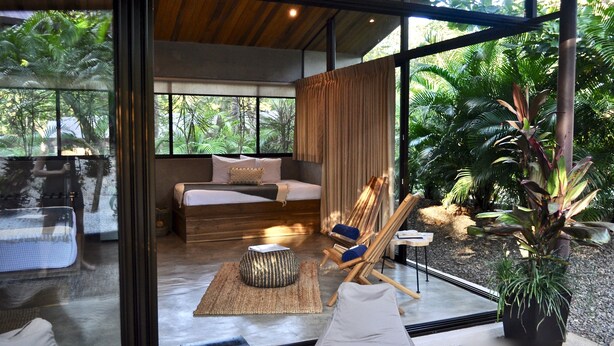 Een moderne kamer met een bed, een zithoek met een fauteuil en poef, en een groot raam met uitzicht op een tuin.
