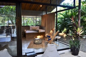 Ein modernes Zimmer mit einem Bett, einer Sitzecke mit Sessel und Hocker sowie einem großen Fenster mit Blick auf einen Garten.