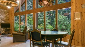 L’intérieur est en bois et comprend une table à manger, des chaises et une cheminée. De grandes fenêtres offrent une vue sur la forêt environnante.