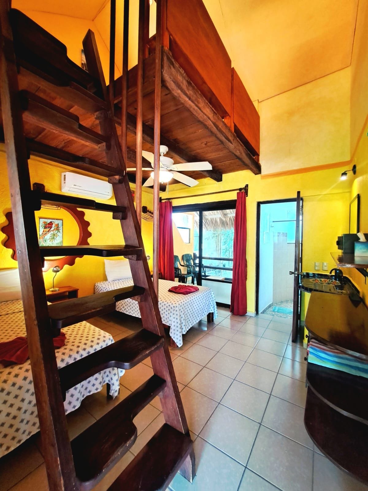 Ein Zimmer mit einer Holztreppe, zwei Betten mit gemusterten Bettbezügen, einem Deckenventilator und einem Fenster mit Vorhängen.