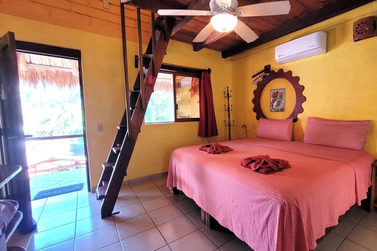 A bedroom with a bed, a staircase, an air conditioning unit, a wooden ceiling, and a window.