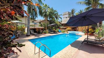 A swimming pool with lounge chairs and umbrellas, surrounded by palm trees and a building.