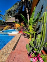 Uma área de piscina com uma estrutura de telhado de palha, cercada por plantas tropicais e um cacto.