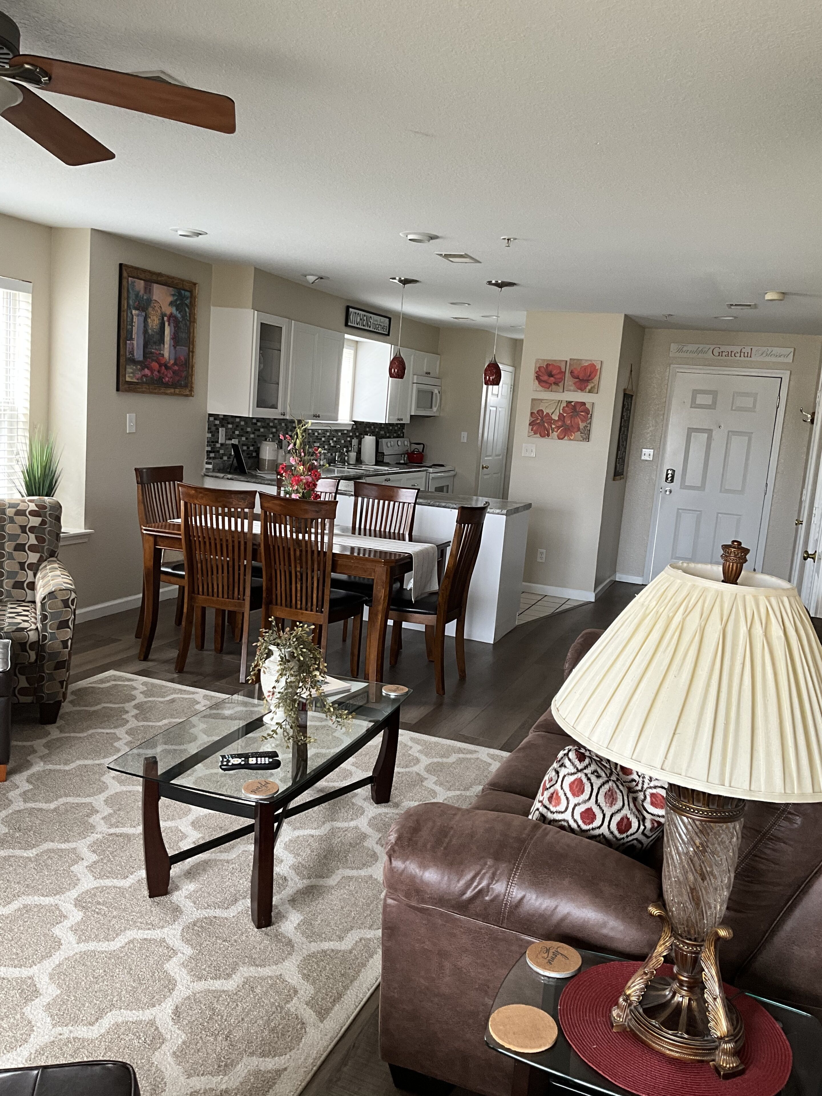 A living room with a brown leather sofa, a glass coffee table, and a dining area with wooden chairs and a kitchen in the background.
