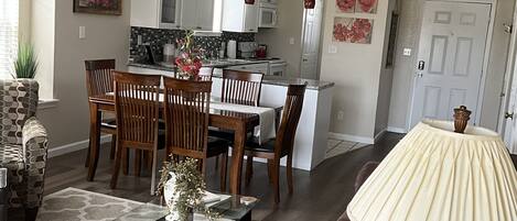 A living room with a brown leather sofa, a glass coffee table, and a dining area with wooden chairs and a kitchen in the background.