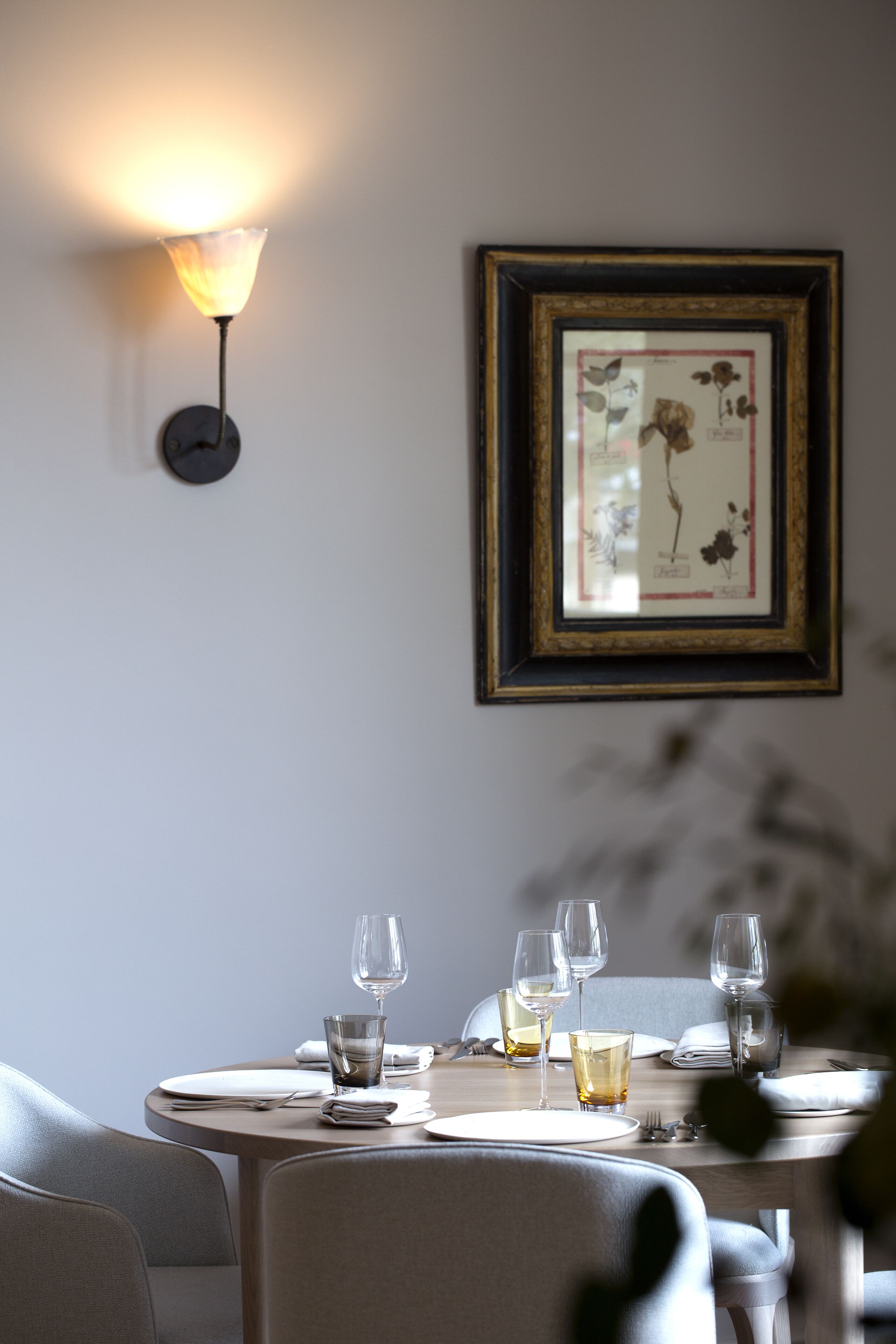Una mesa de comedor dispuesta con platos, vasos y cubiertos, una ilustración botánica enmarcada en la pared y una lámpara fijada a la pared.