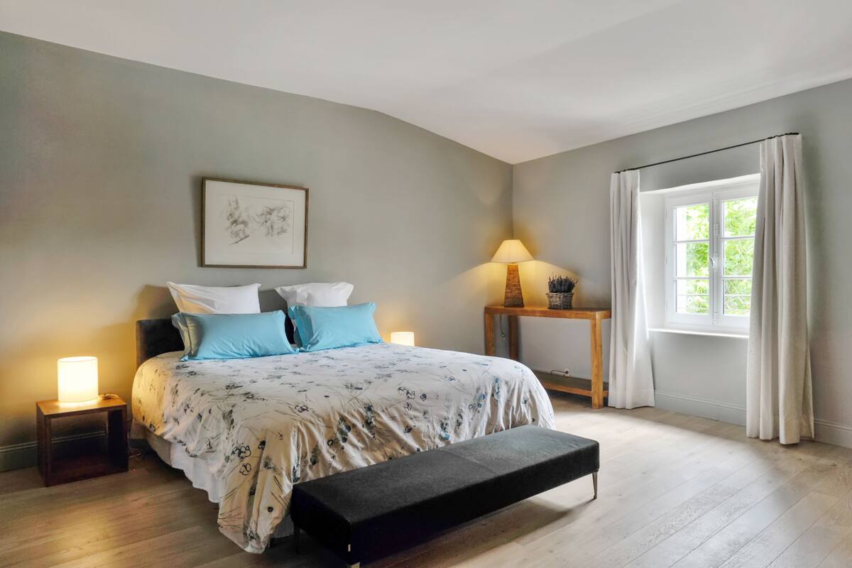 A bedroom with a bed, bedside tables, a bench, a window with curtains, and a wall-mounted picture.
