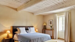 Una camera da letto con soffitto in legno, un letto con testiera, lampade sui comodini, un comodino con una lampada, una finestra con tende e una panca in legno.