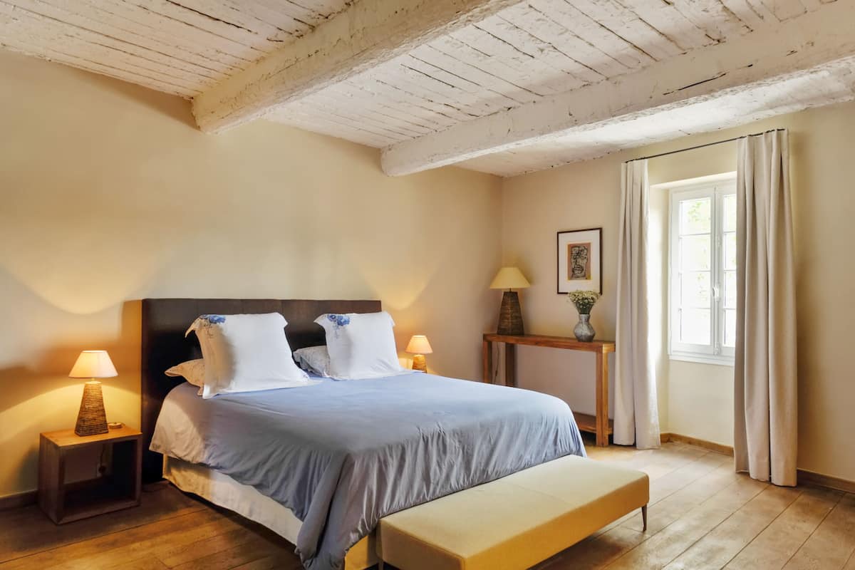 A bedroom with a wooden ceiling, a bed with a headboard, bedside lamps, a bedside table with a lamp, a window with curtains, and a wooden bench.