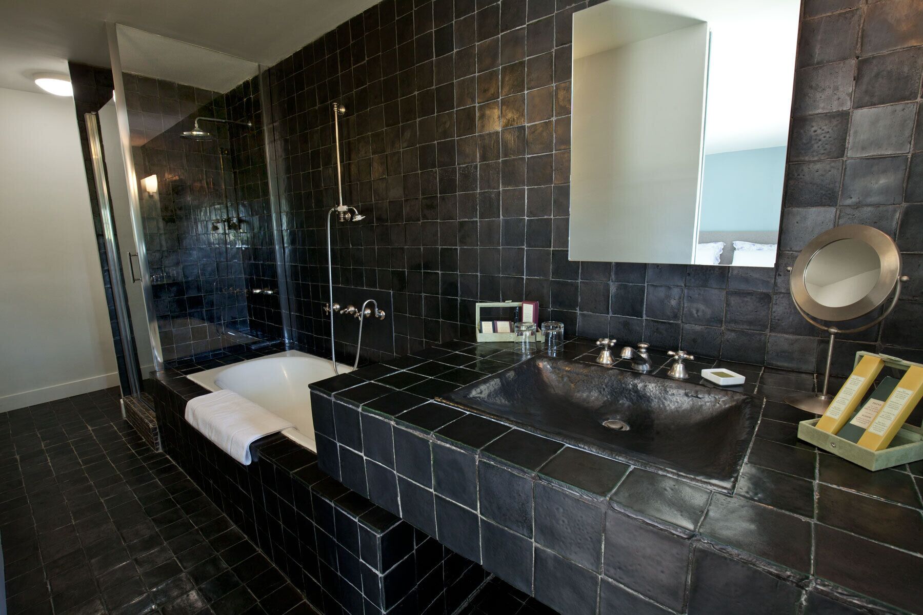 Un baño moderno con pared de azulejos negros, ducha con cerramiento de vidrio, bañera blanca y lavamanos con espejo.