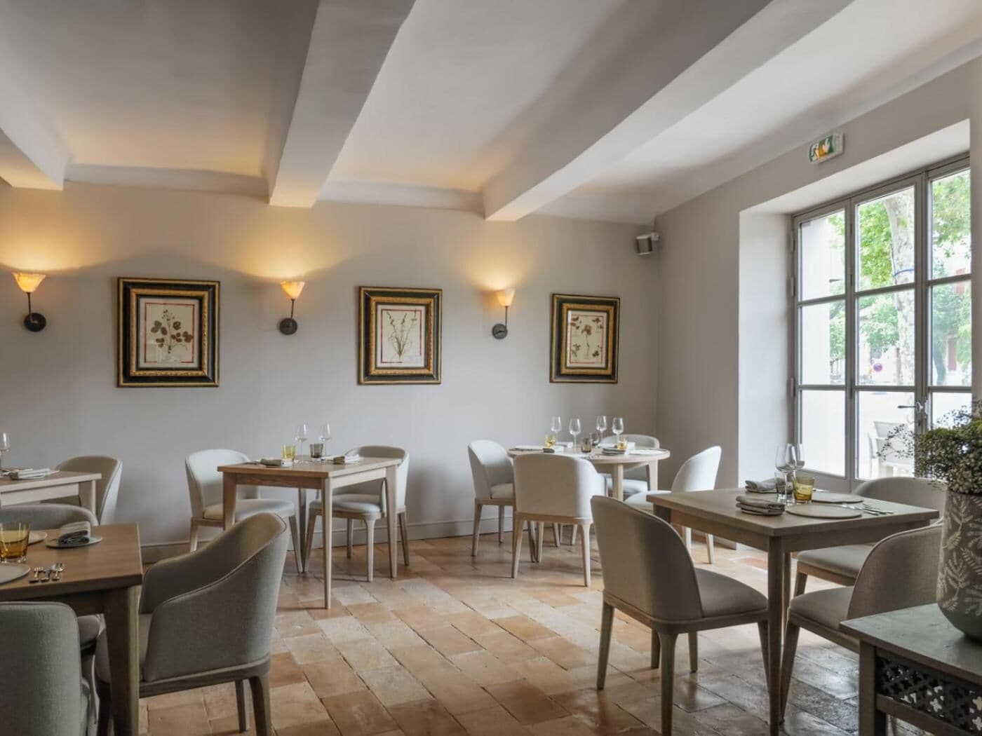 Un espace repas comprenant plusieurs tables et chaises, des œuvres d’art encadrées aux murs et de grandes fenêtres laissant entrer la lumière naturelle.