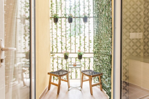 A room with two wicker chairs, a small table, and a view through a window with hanging plants.