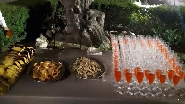 An outdoor dining setup with a table set for a meal, including bowls of food and a row of glasses filled with a red beverage.