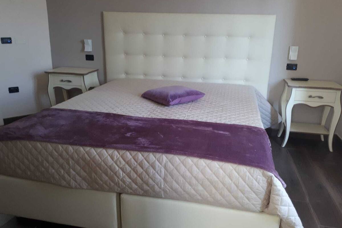 A neatly made bed with a quilted bedspread and a single purple pillow, flanked by two bedside tables with lamps.