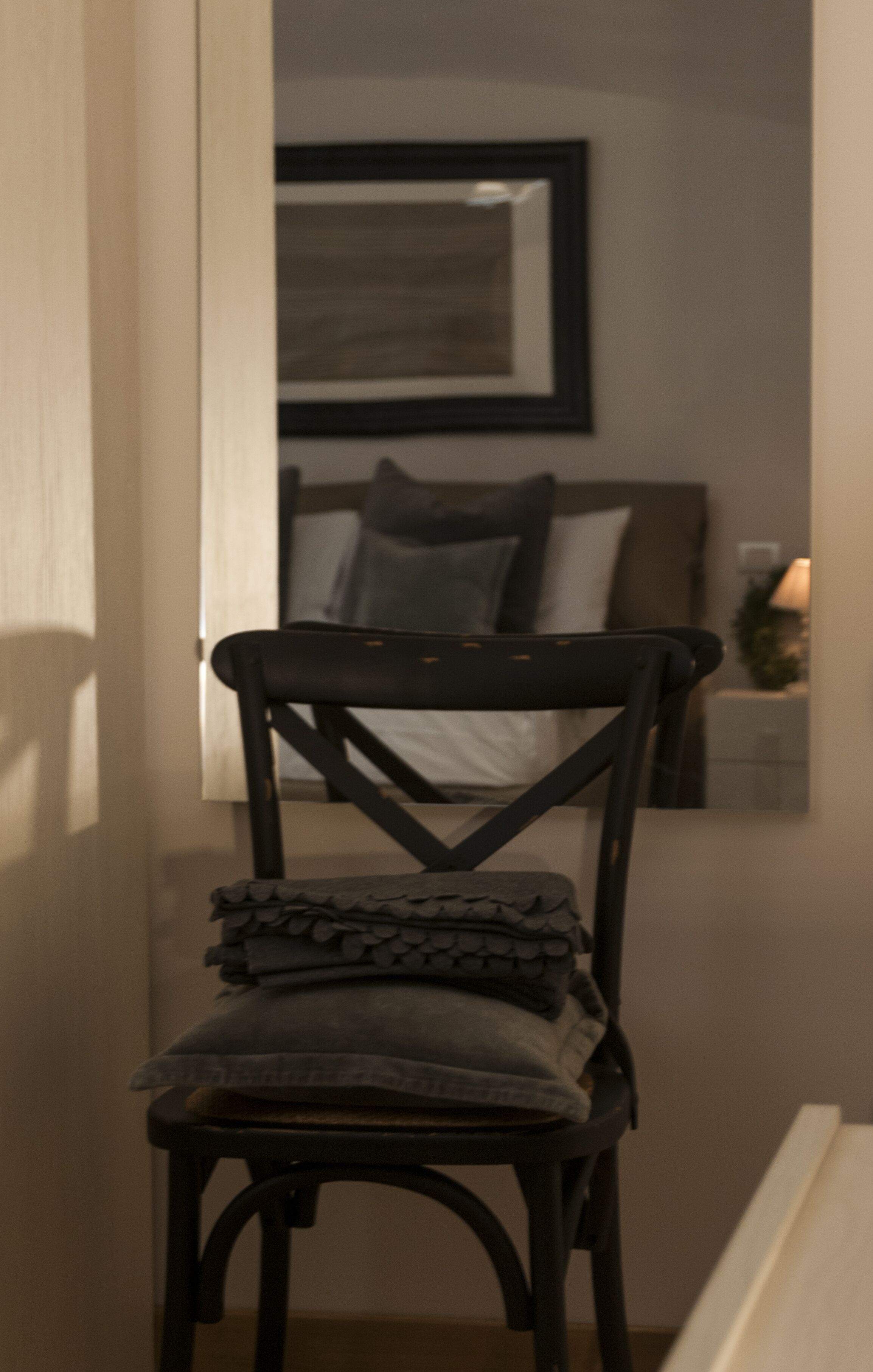 Une chaise dotée d’une structure sombre et d’un coussin, placée dans un couloir où un miroir reflète un lit et des oreillers.