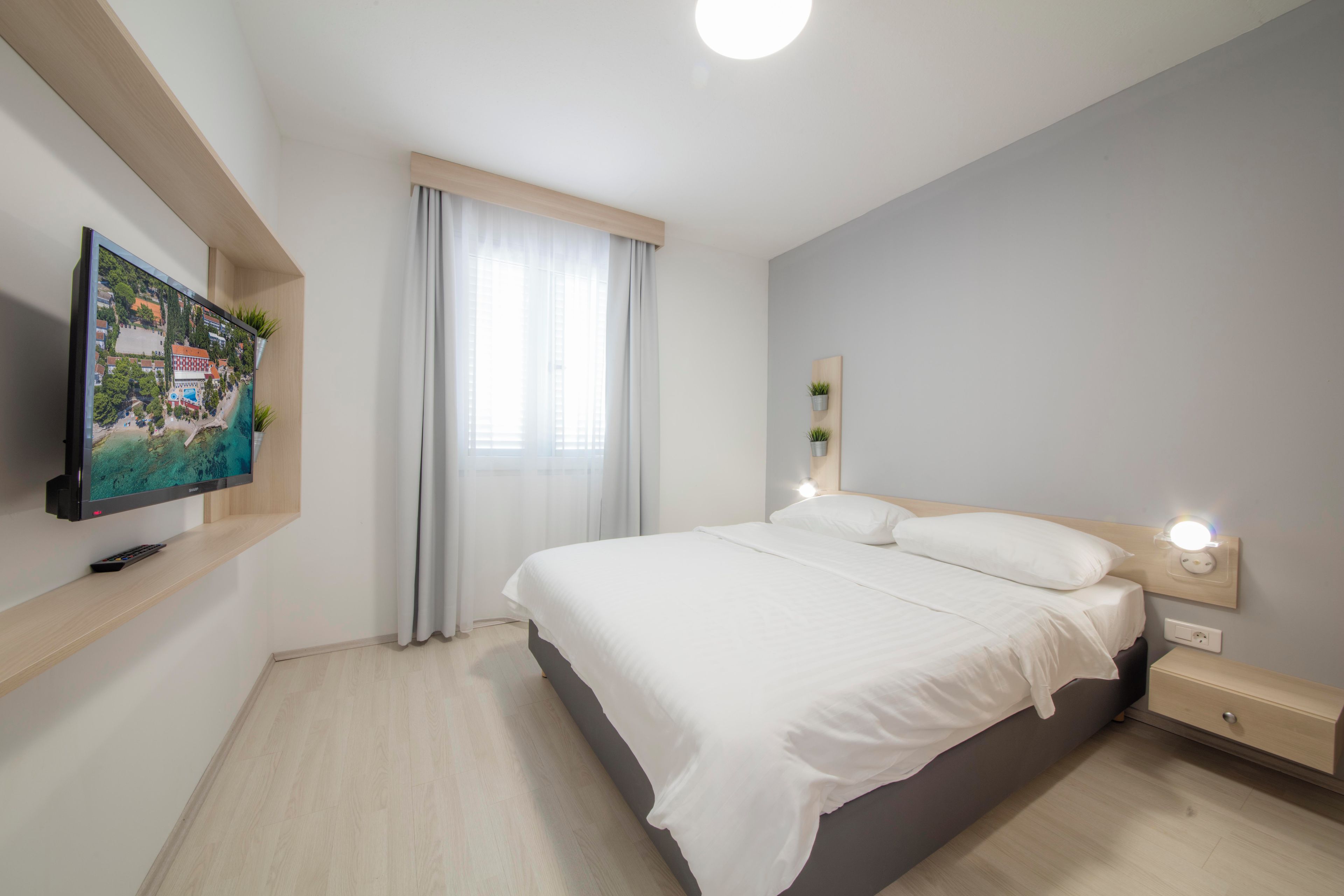 Un dormitorio moderno con cama, mesitas de noche, televisor montado en la pared y ventana con cortinas.