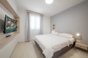 Een moderne slaapkamer met een bed, nachtkastjes, een aan de muur bevestigde televisie en een raam met gordijnen.