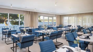 A modern dining area with blue chairs and tables, large windows with curtains, and a view of the sea.