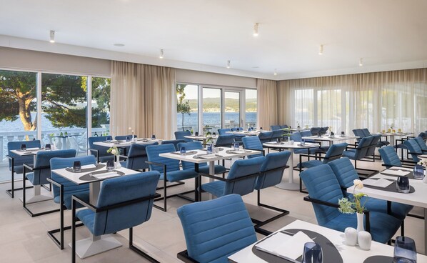 A modern dining area with blue chairs and tables, large windows with curtains, and a view of the sea.