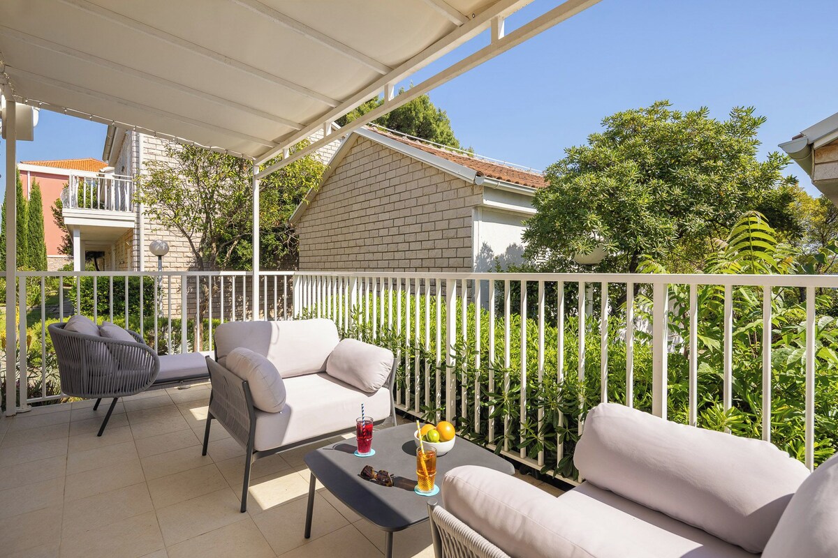 A covered outdoor seating area with a white railing, a table with drinks, and a view of a house and trees.
