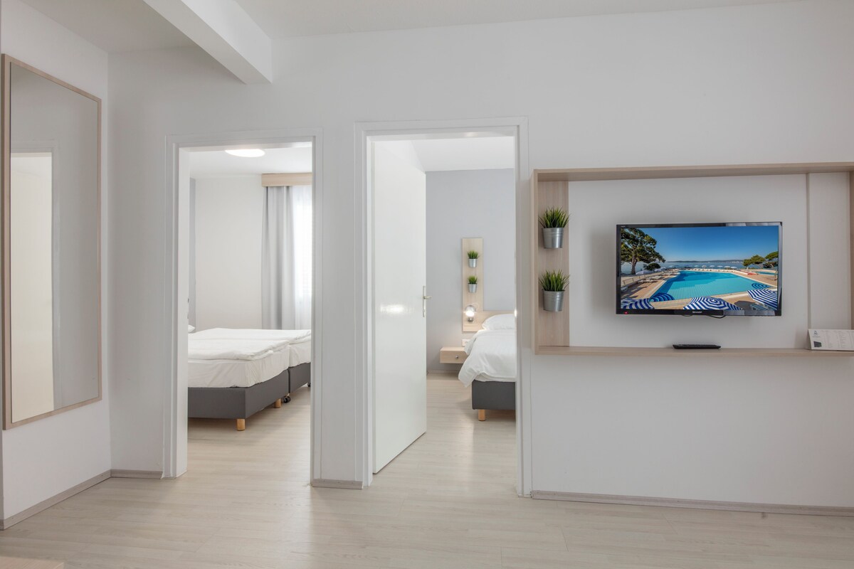 A modern hotel room with a large TV displaying a beach view, a bed, a bedside table, and a small plant.