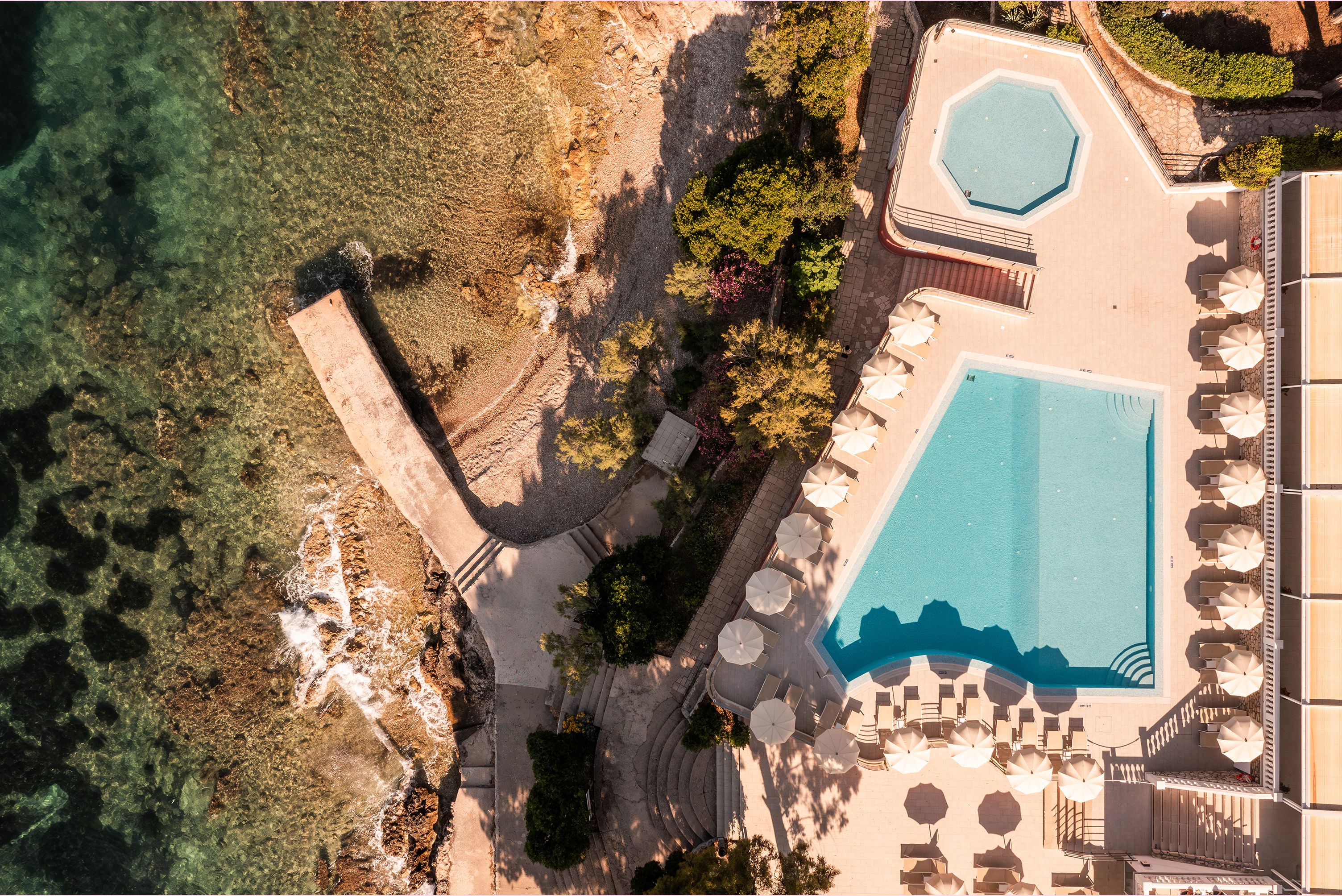 An aerial view of a resort with a large swimming pool, sun loungers, and a landscaped area with trees and a stone pathway.