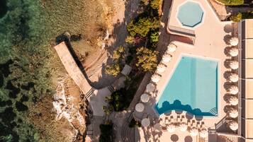 An aerial view of a resort with a large swimming pool, sun loungers, and a landscaped area with trees and a stone pathway.