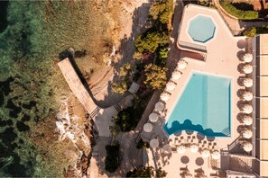 An aerial view of a resort with a large swimming pool, sun loungers, and a landscaped area with trees and a stone pathway.
