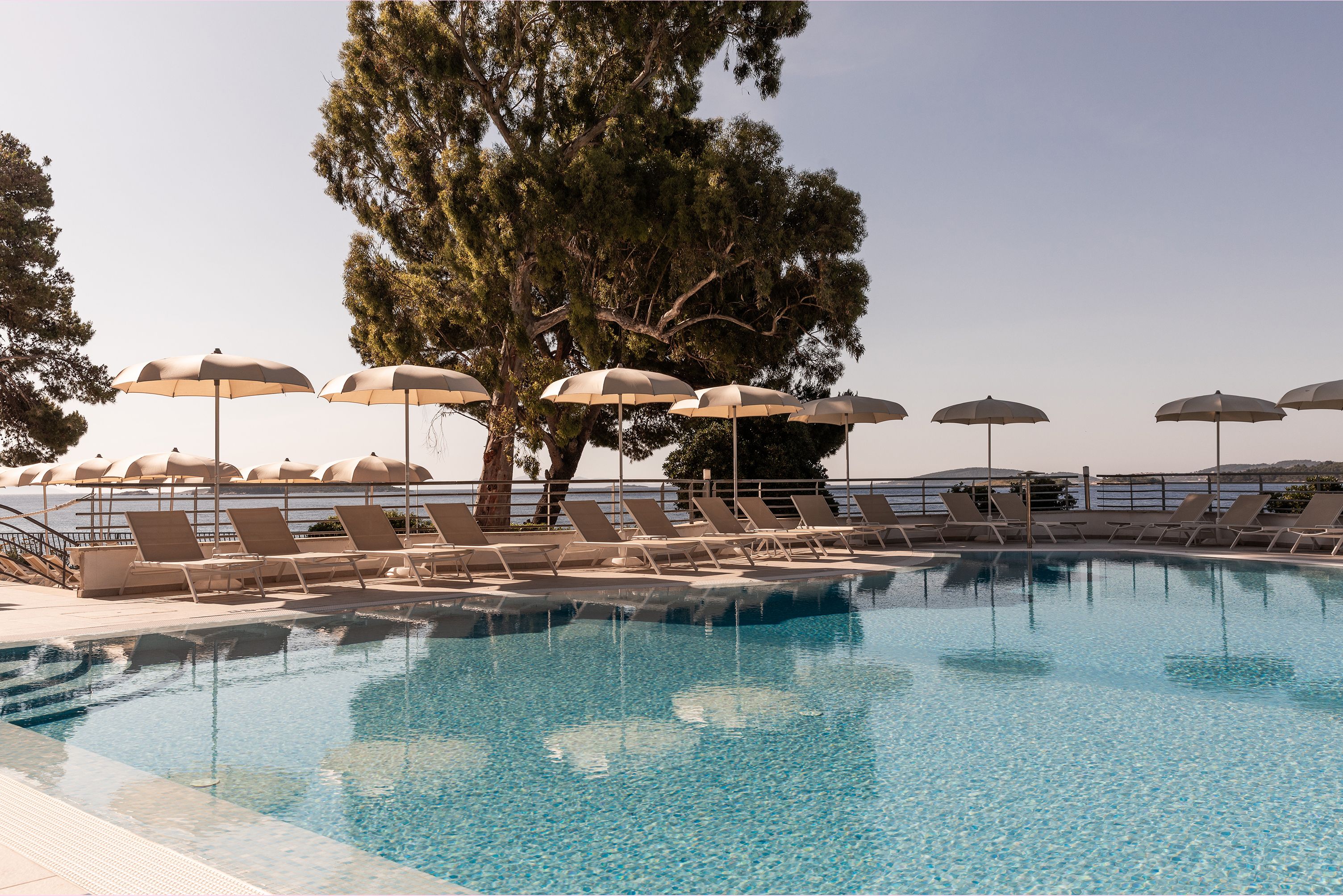 A swimming pool with lounge chairs and umbrellas, surrounded by trees and a view of the sea.